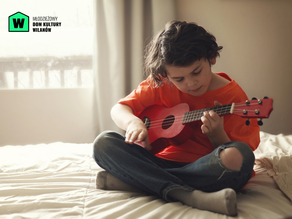 Mały chłopczyk trzymający w ręku czerwone ukulele, uczy się na nim grać.